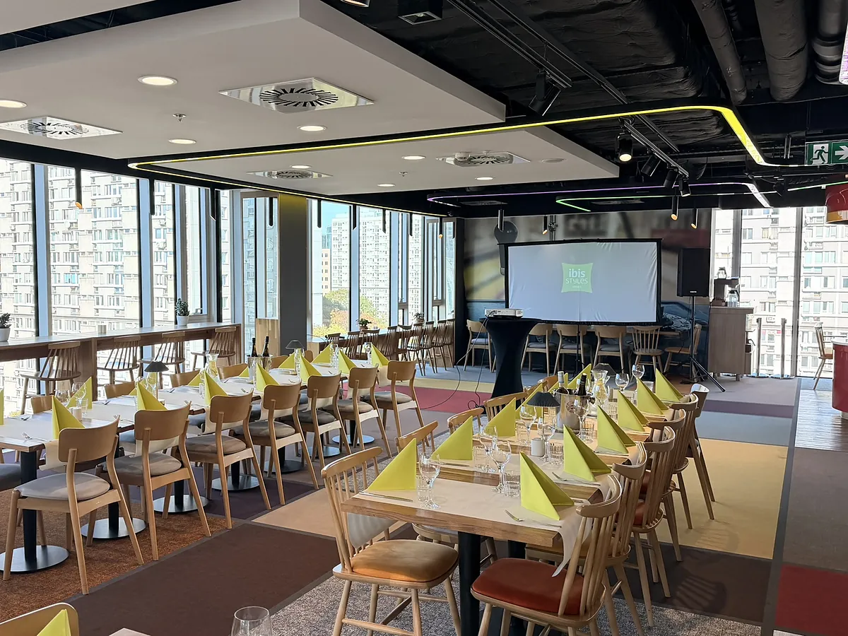 Restauracja na 7. piętrze przygotowana na kolację z prezentacją — nakryte stoły z żółtymi serwetkami, ekran z logo ibis Styles, rzutnik i panoramiczne okna z widokiem na miasto