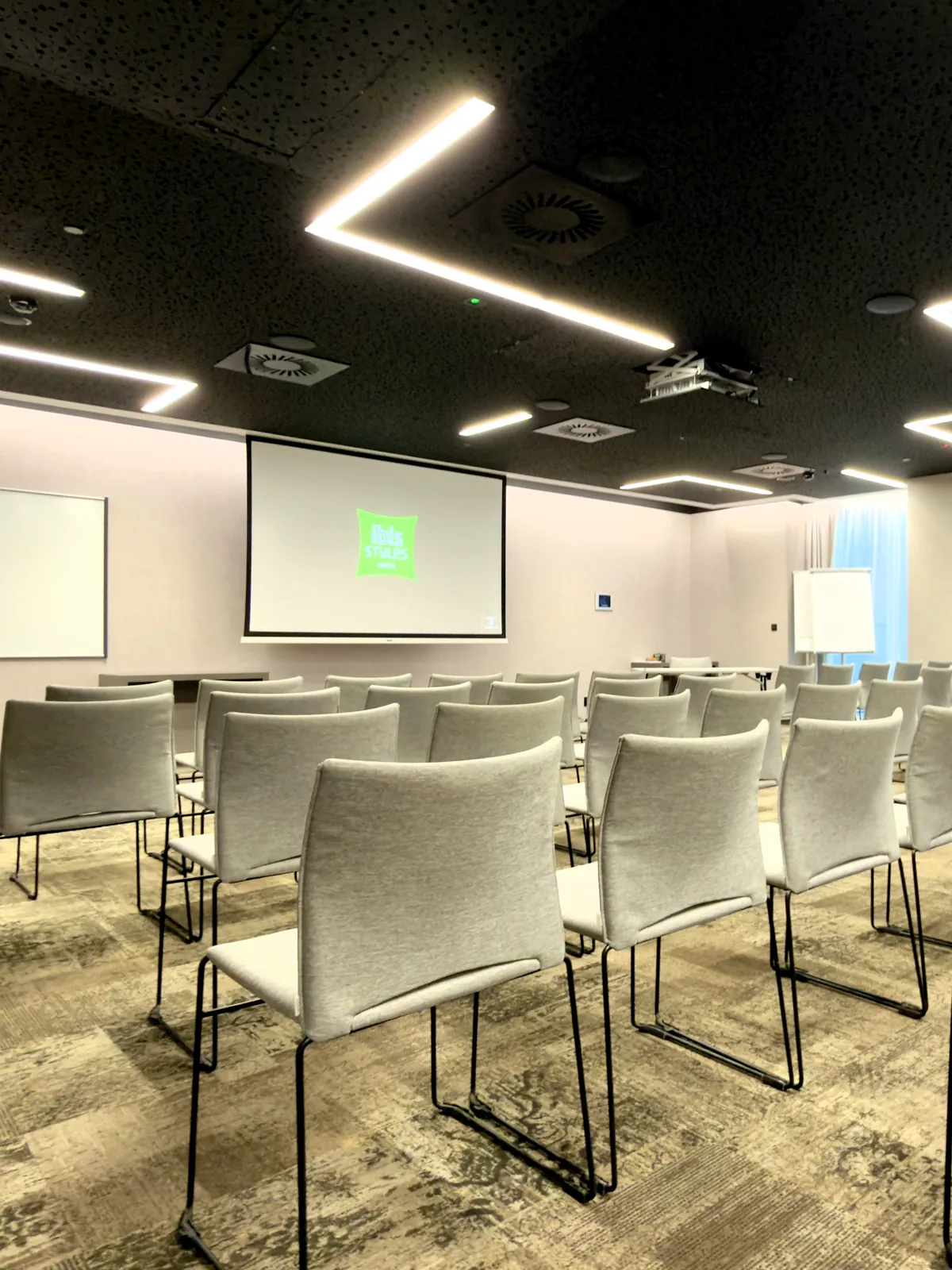 Meeting room at ibis Styles Warszawa City in theatre layout — rows of chairs, a projection screen with the ibis Styles logo, a flipchart and windows with natural light