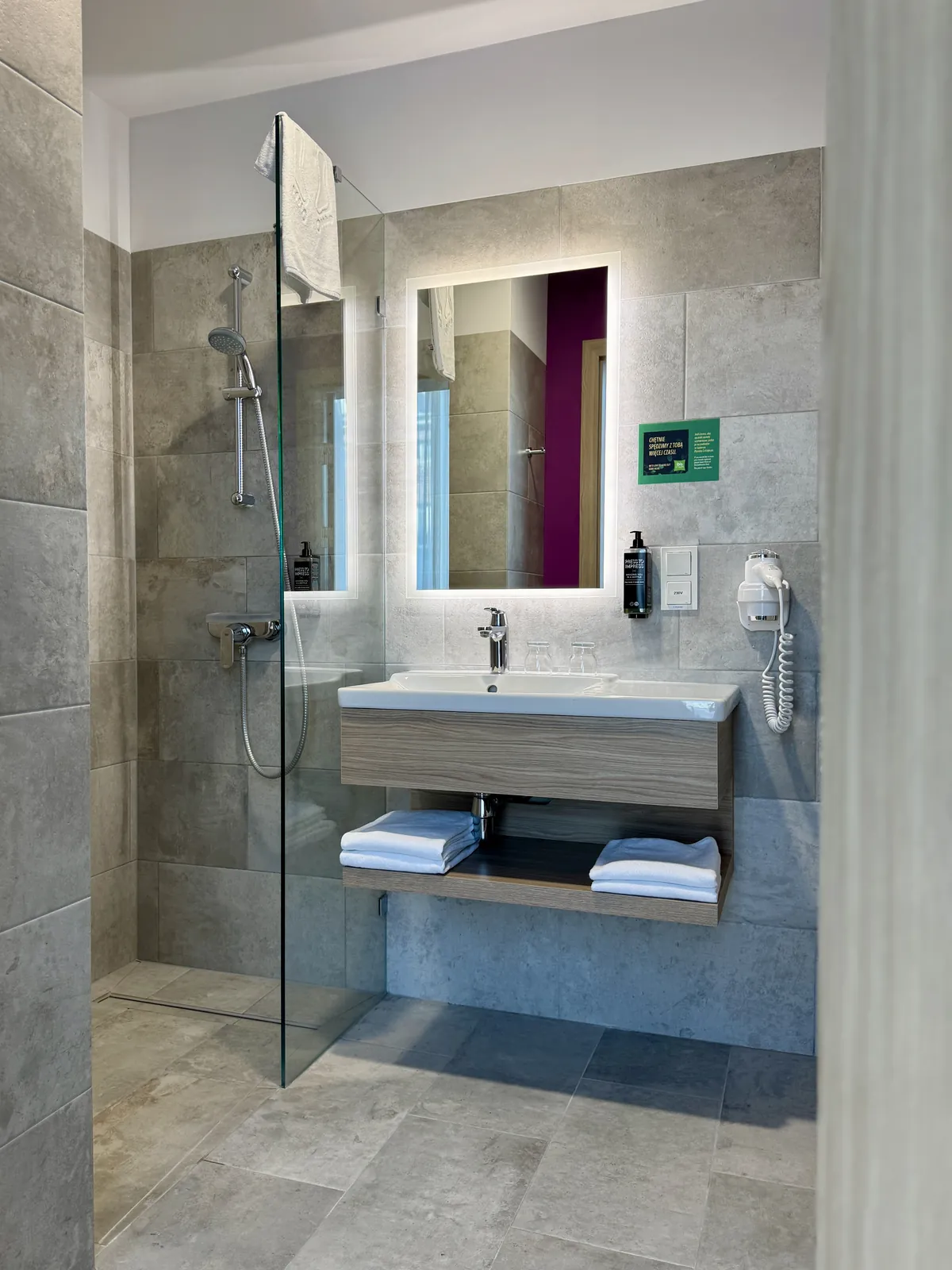 Bathroom in the room — walk-in shower behind a glass panel, washbasin with a backlit mirror and towels on a shelf