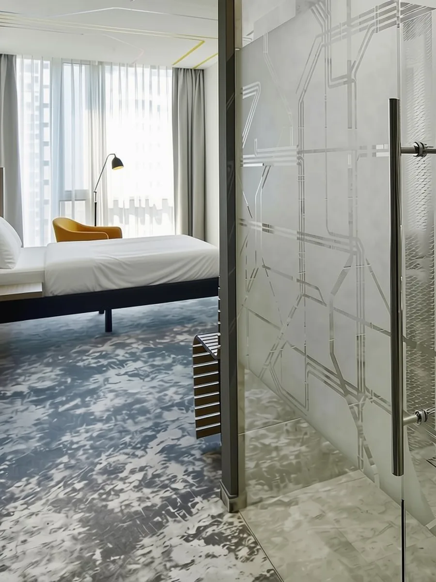 Standard room at ibis Styles Warszawa City — bed, yellow armchair, panoramic window and a glass bathroom wall etched with a map of Warsaw tram lines