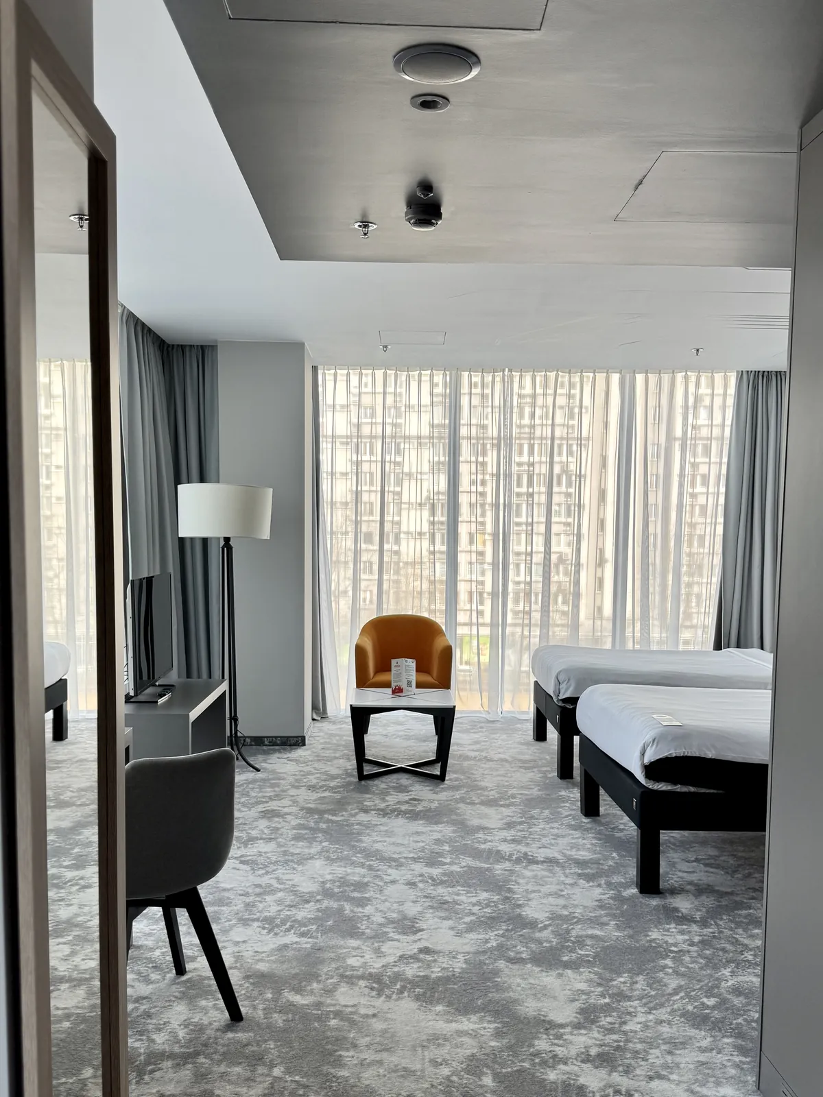 Twin room — two separate beds, an orange armchair, a floor lamp, a mirror and a floor-to-ceiling window overlooking the city