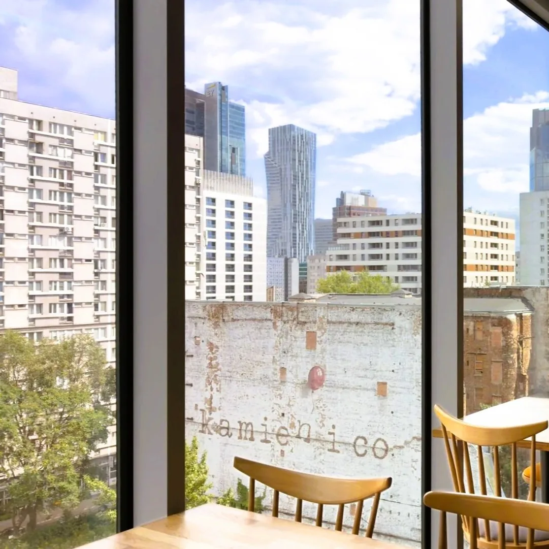 Seventh-floor breakfast room at ibis Styles Warszawa City — floor-to-ceiling panoramic windows, tables by the glass and a view over central Warsaw and a pre-war tenement house on Waliców Street
