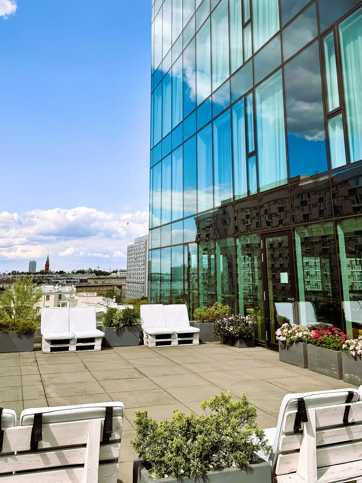 Seventh-floor terrace — pallet loungers, planters with flowers, the hotel's glass façade reflecting clouds and the Warsaw panorama beyond